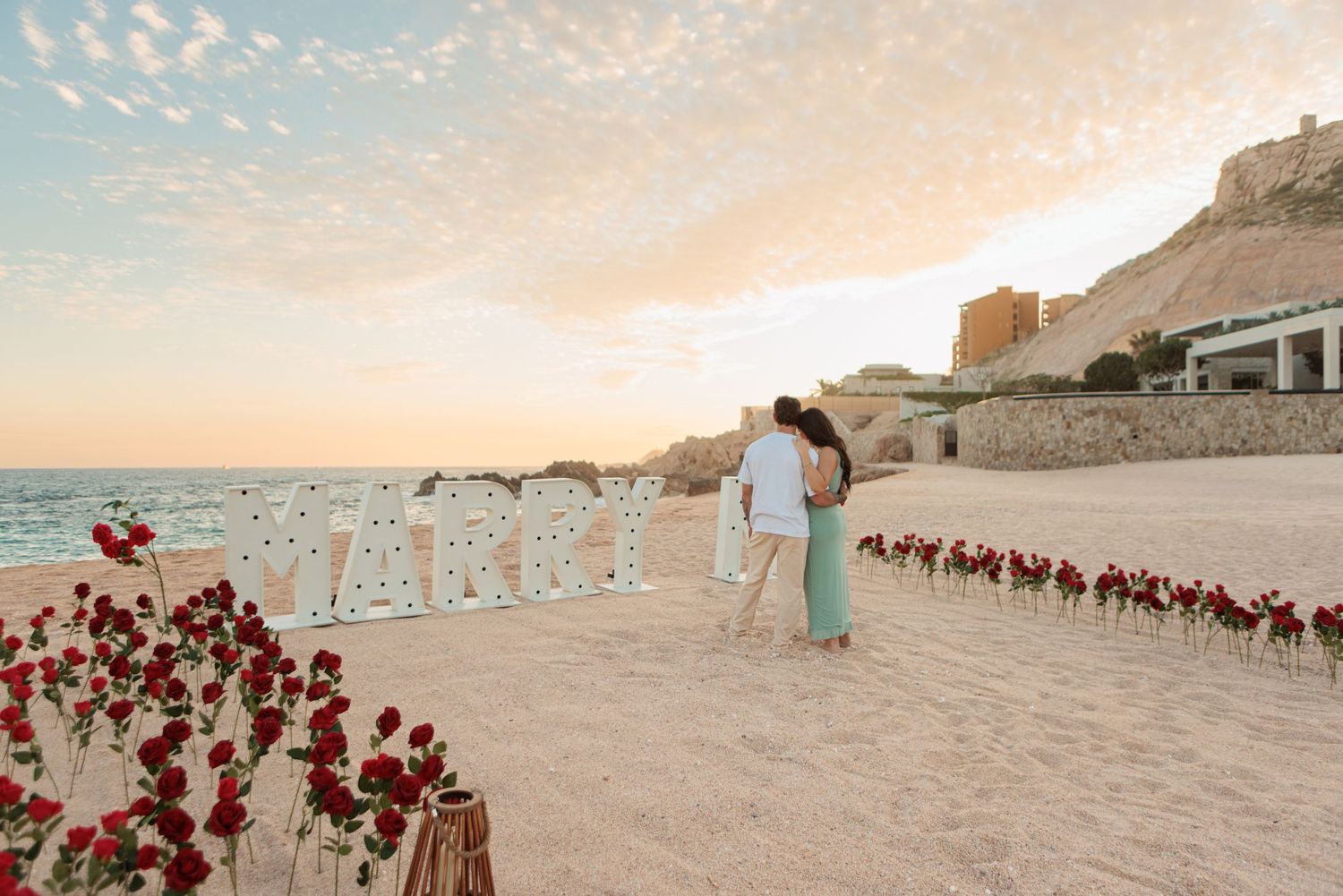 Romantic beach proposal setup with MARRY ME letters and red roses at sunset.