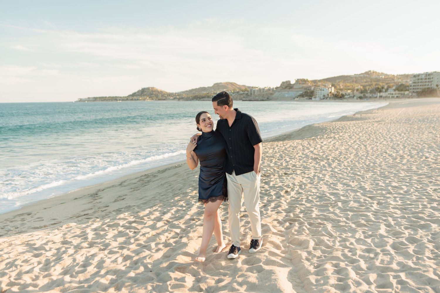 Couple holding hands and smiling on a sandy beach with turquoise ocean waves and rocky hills in the background.