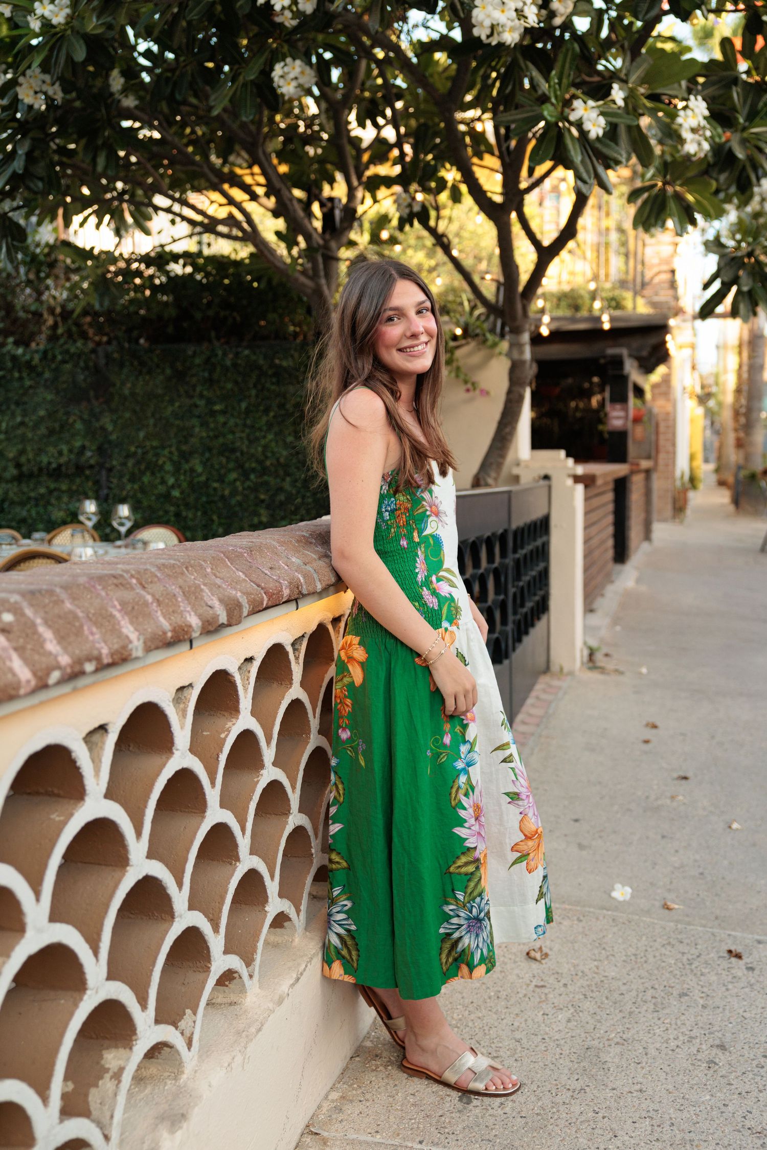 Woman in green floral midi dress smiling outdoors near decorative wall with blooming trees.