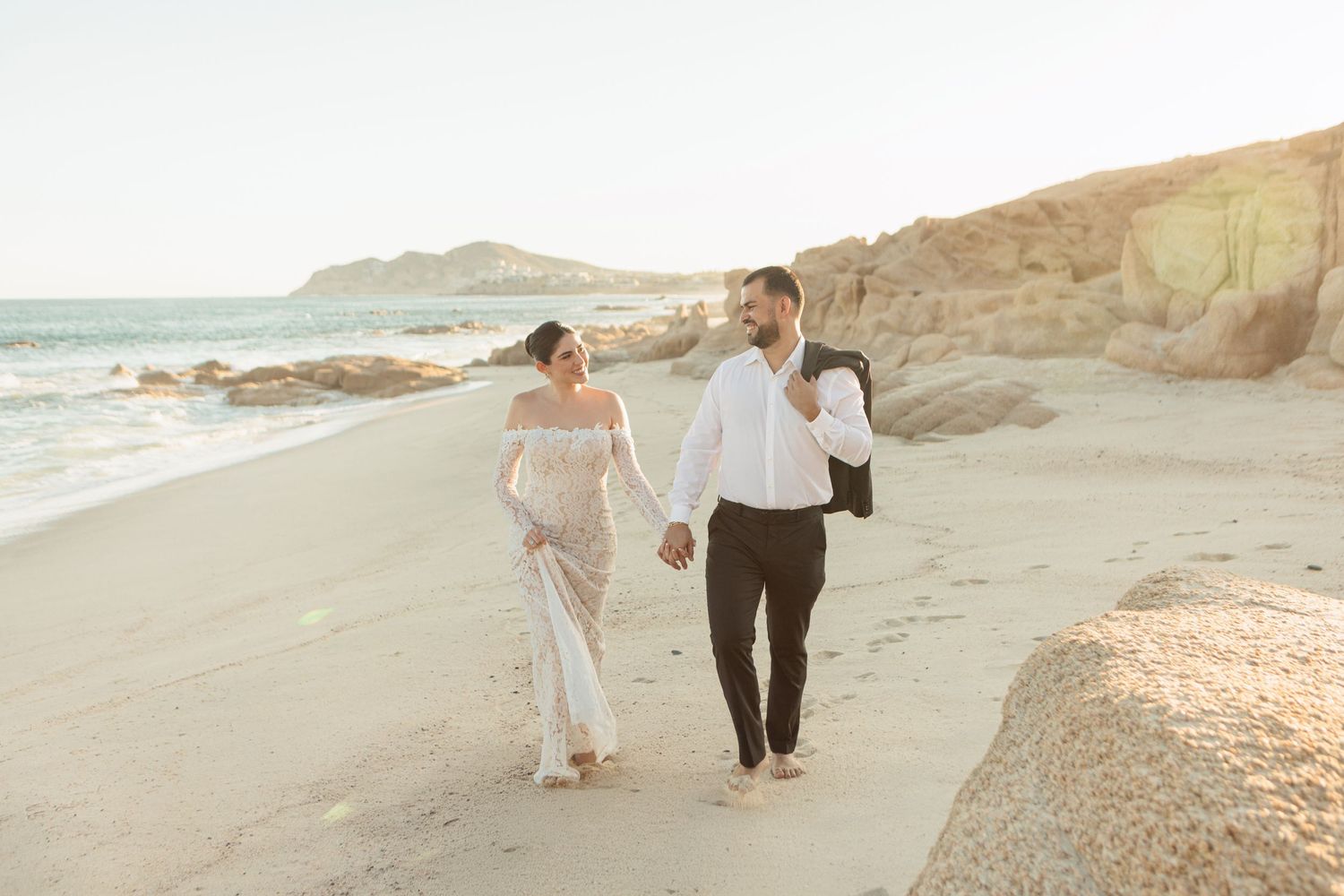 Bride and groom walk hand-in-hand along sunlit beach, groom carrying his jacket over his shoulder.