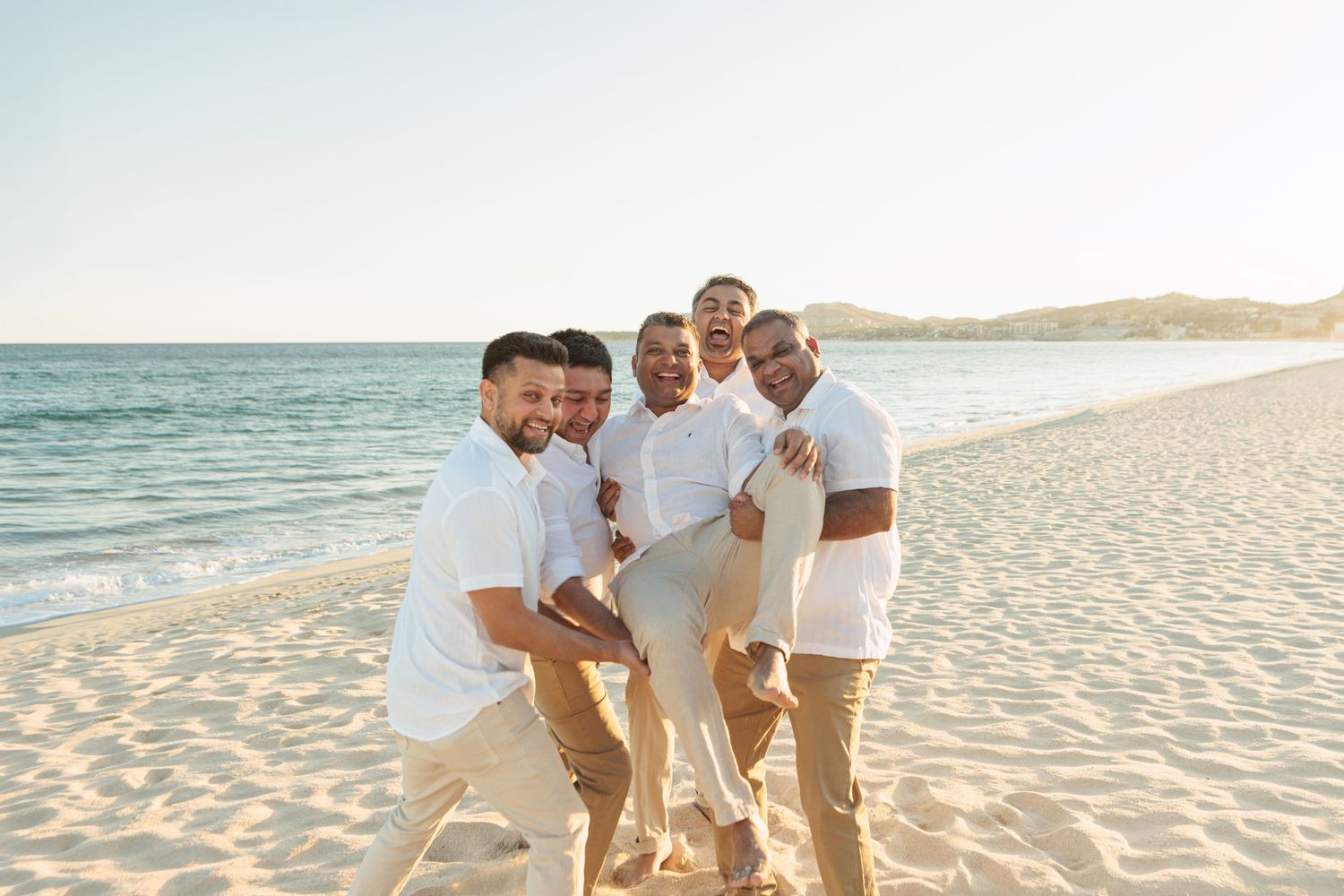 Five men in white shirts laughing and playing on sunny sandy beach near ocean shoreline.