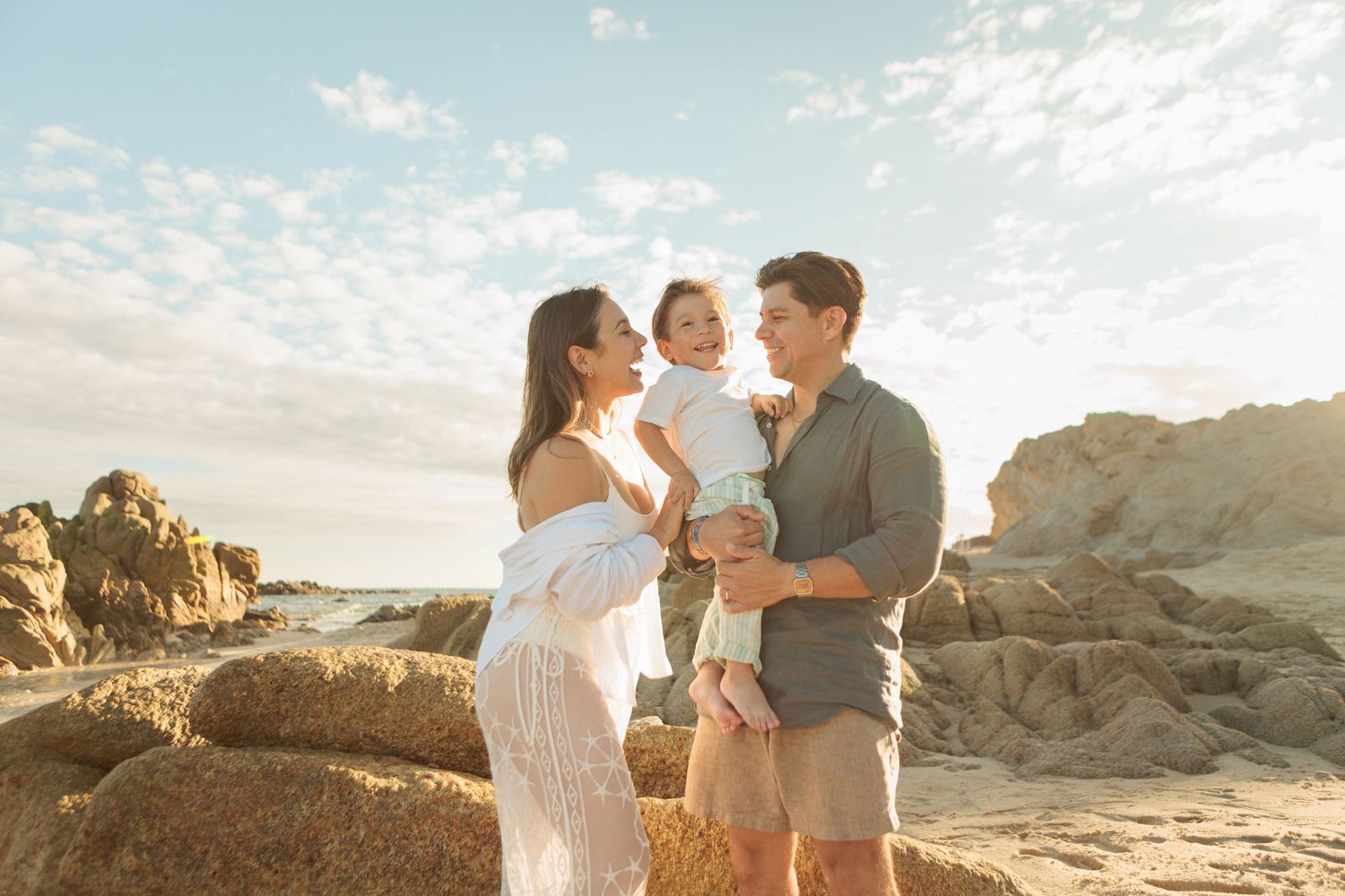 Smiling family of three posing together on a sunny beach with rocky formations and blue sky behind them.