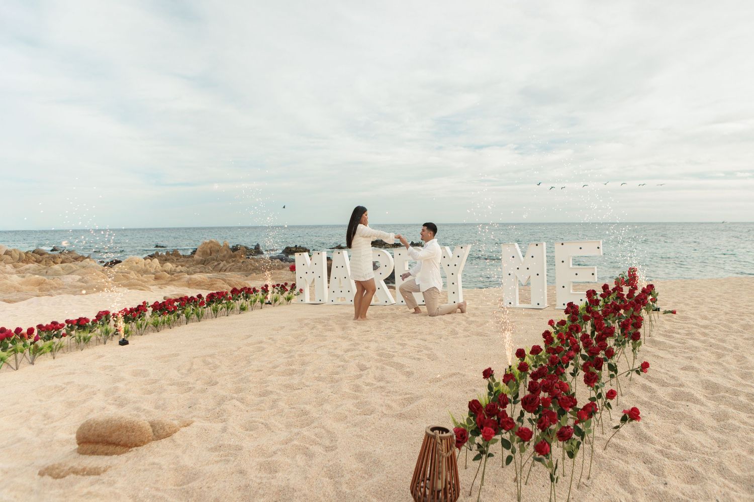 Beach proposal setup with red roses and lit marquee letters spelling MARRY ME.