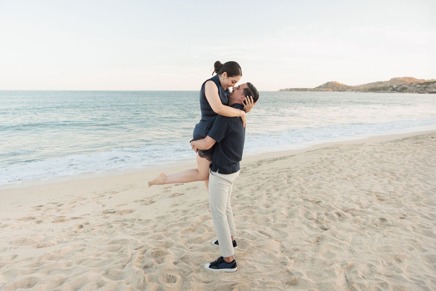 Couple sharing a romantic kiss on a sandy beach, man lifting woman in his arms with ocean waves in the background.