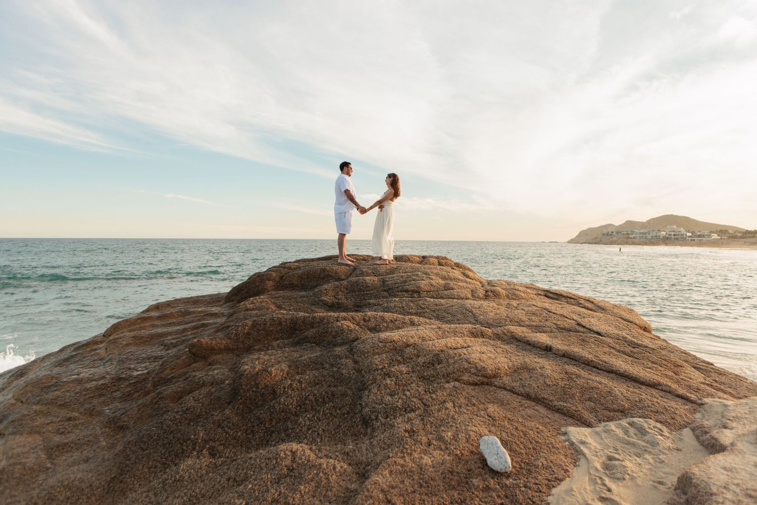 Couple holding hands atop a large rock with ocean and mountain views at sunset.
