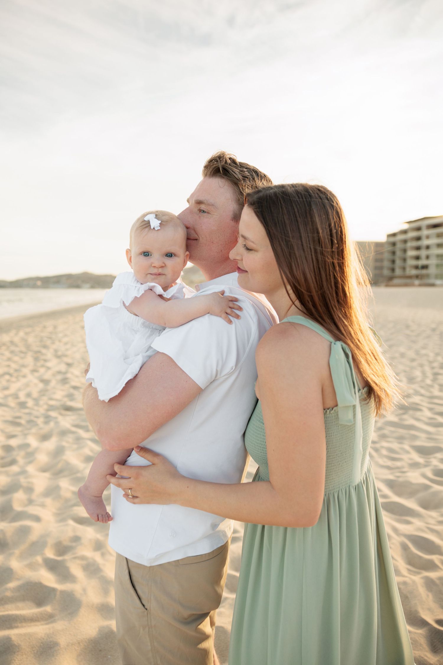A couple stands on a sandy beach, the man kissing their baby girl while the woman in a green dress looks on lovingly.