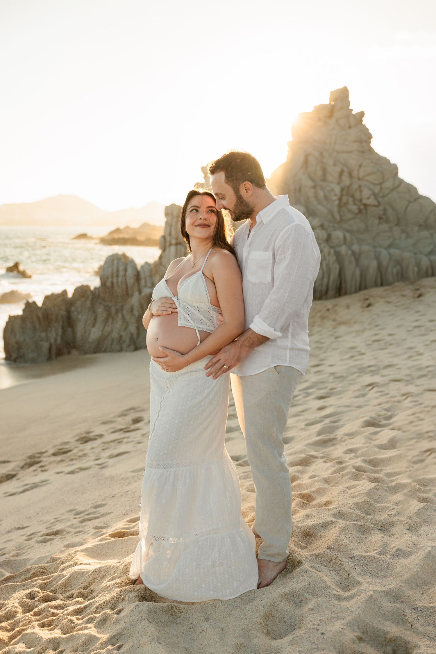 Couple embracing on sandy beach at sunset, pregnant woman in white dress with rocky backdrop.