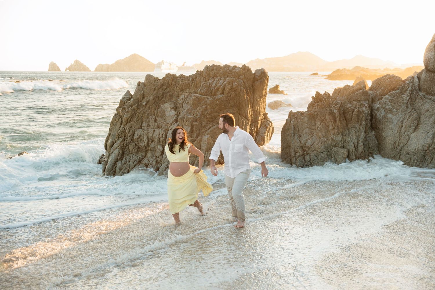 Happy couple running hand in hand along rocky beach shore during beautiful golden sunset.