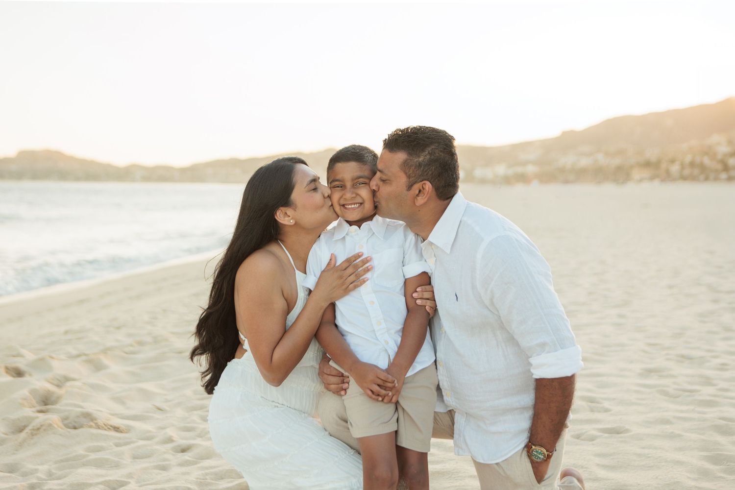 Parents in white outfits kissing their young son on the cheeks on a sunny beach.