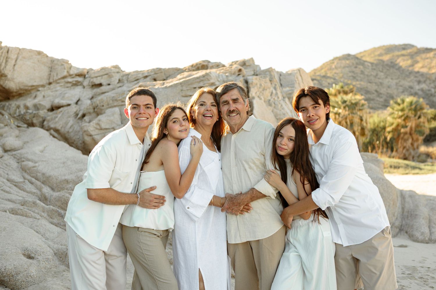 Family of six poses together near rocky formations and palm trees at sunset.