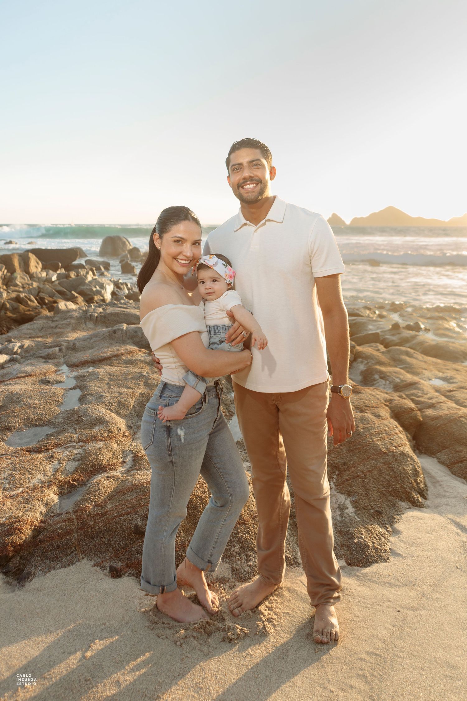 Young family with baby poses on rocky beach at golden hour sunset.