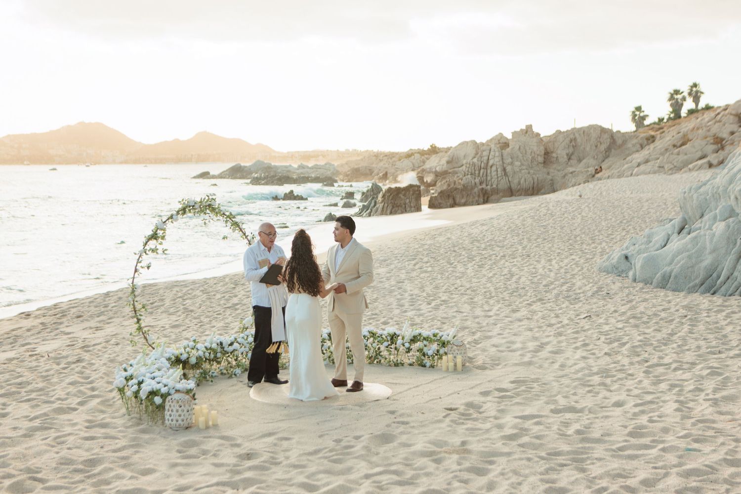 Couple exchanges vows on a sandy beach at sunset with a floral circular arch and ocean backdrop in Cabo San Lucas.