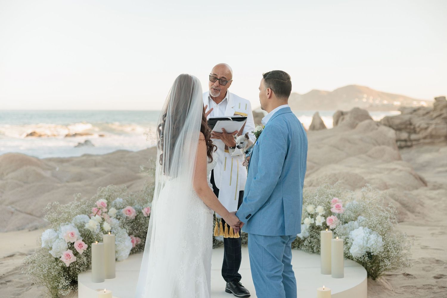 Couple exchanges vows at beach wedding ceremony with officiant, floral arrangements, and rocky ocean backdrop.
