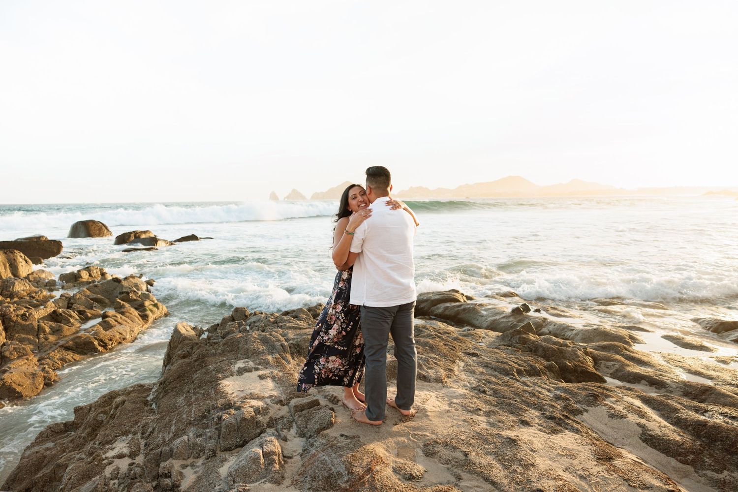 Couple embracing on rocky coastal shore at sunset with ocean waves and mountains in background.