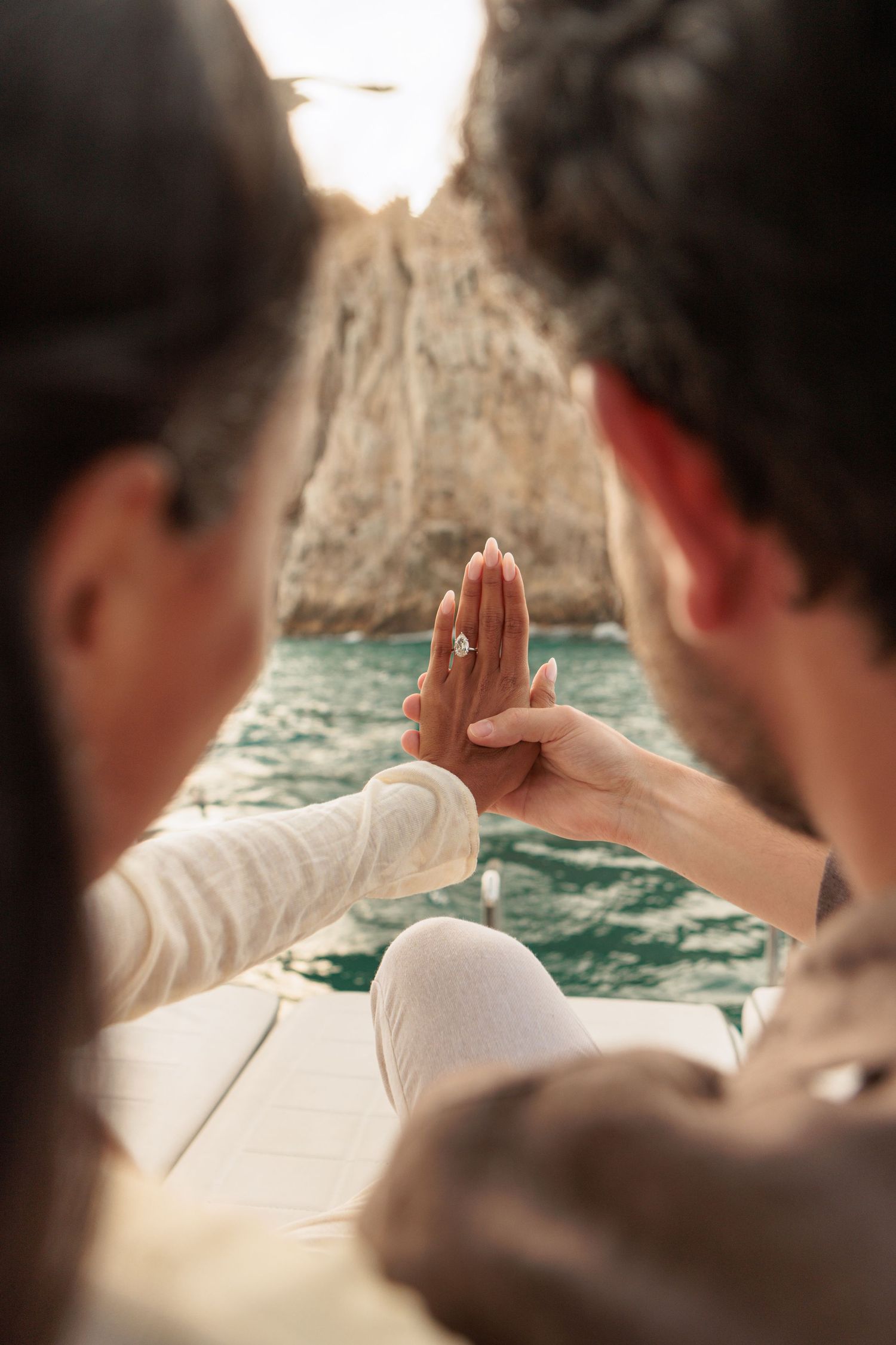 Close-up of couple holding hands showing diamond engagement ring with rocky ocean arch in the background.