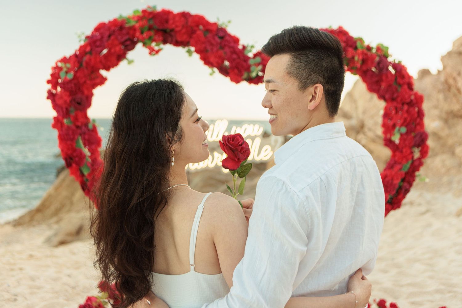 Romantic couple embracing in front of a red rose heart arch on the beach.