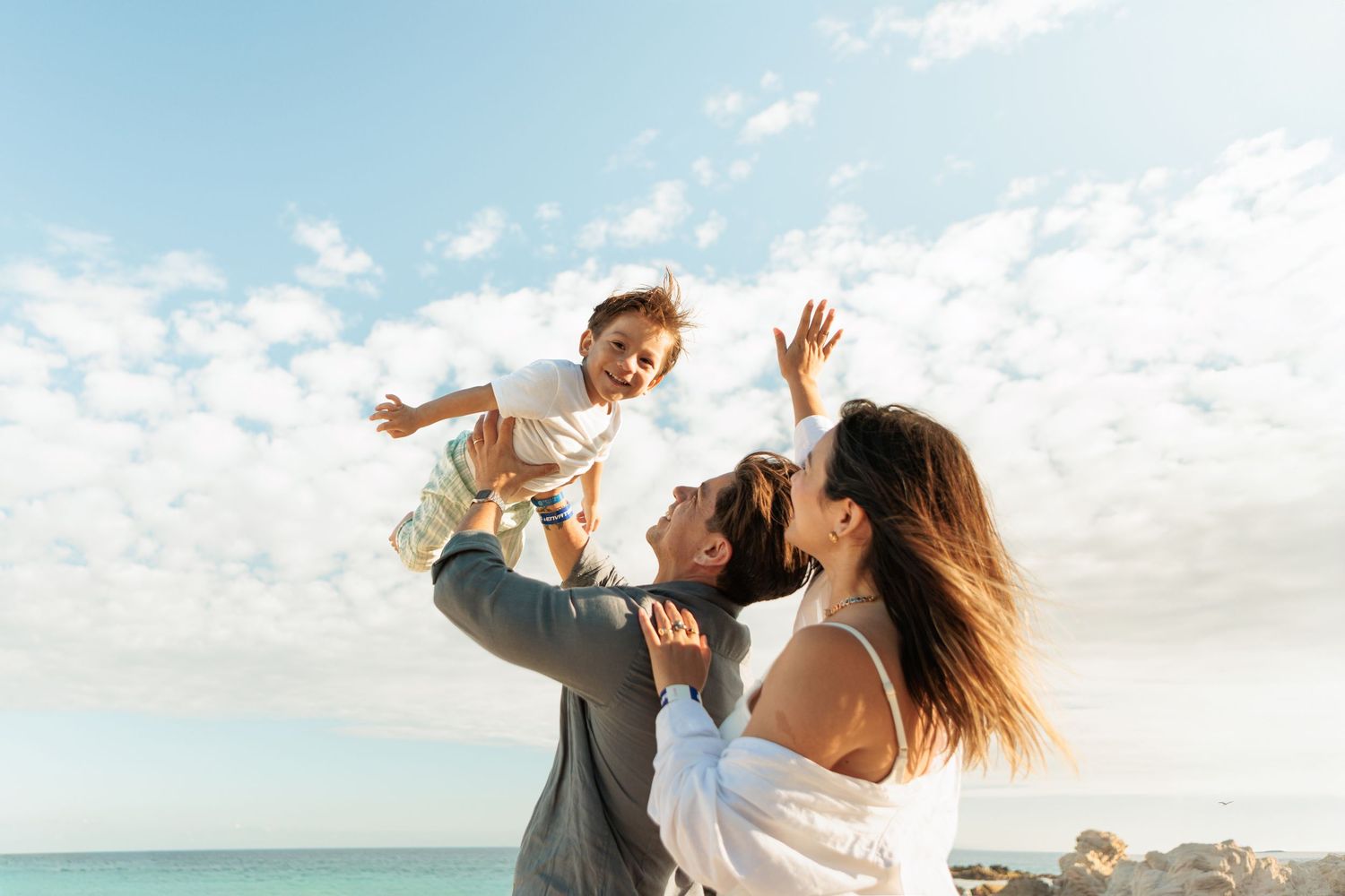 Joyful family at the beach with parents lifting happy child into the blue sky near the ocean.