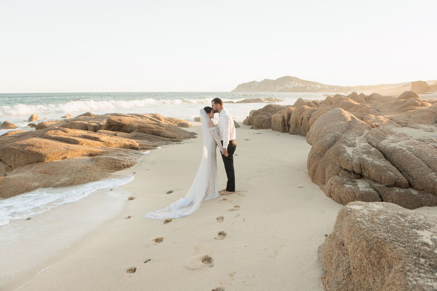 Newlyweds share a kiss on sandy beach with rocky coastline, bride's cathedral veil trailing behind her.