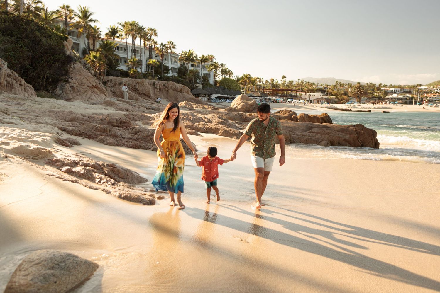 Young family holding hands strolling along sunny beach shoreline with resort in background.