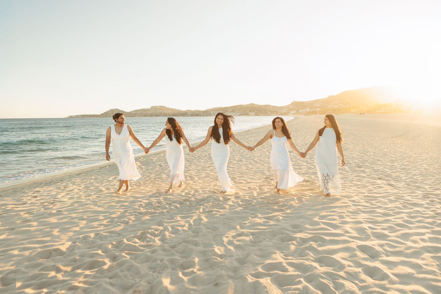 Five women in white dresses hold hands walking on a sunny beach at golden hour.