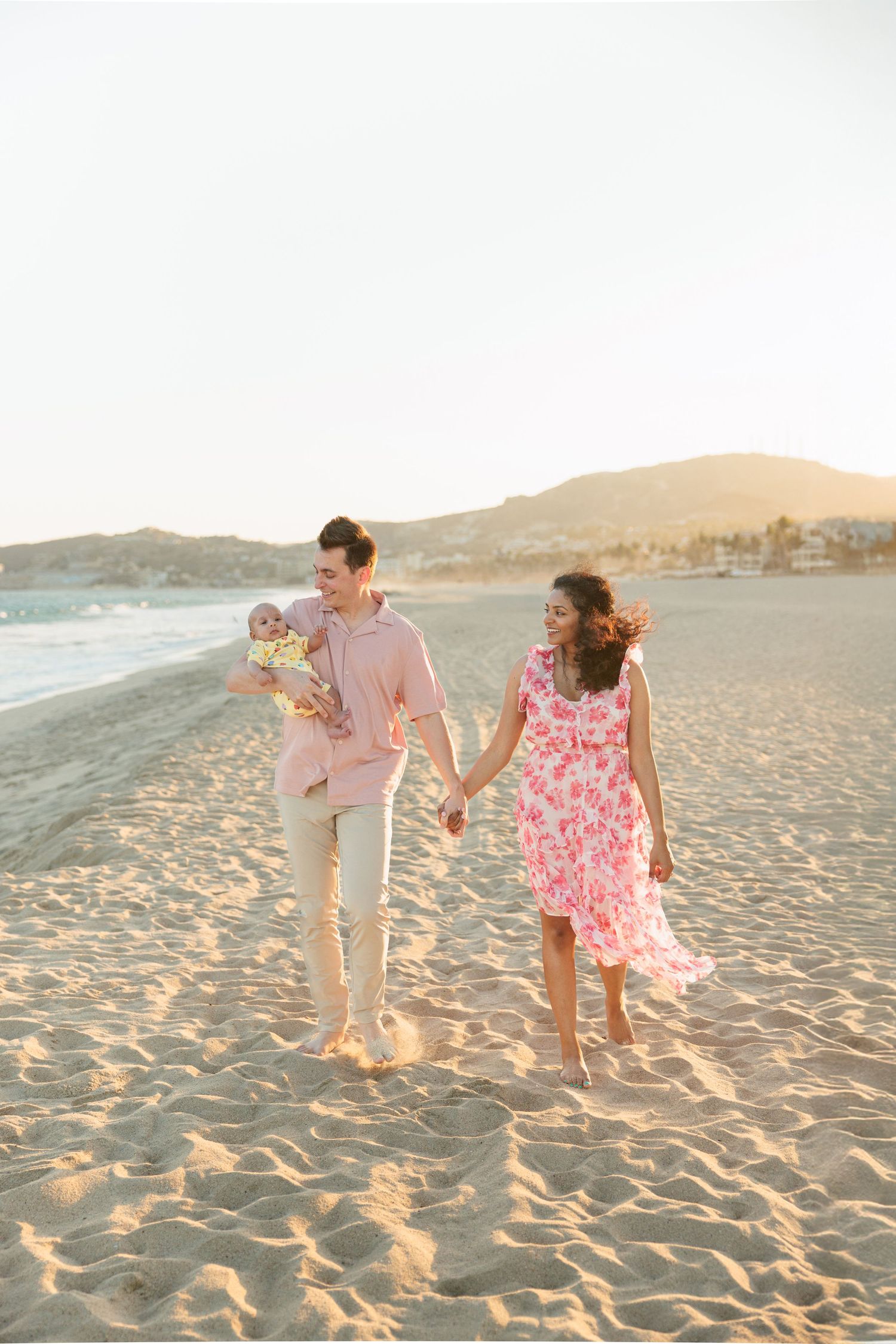 A young family walks hand-in-hand on a sandy beach at sunset, with the man holding a baby in a pink outfit.