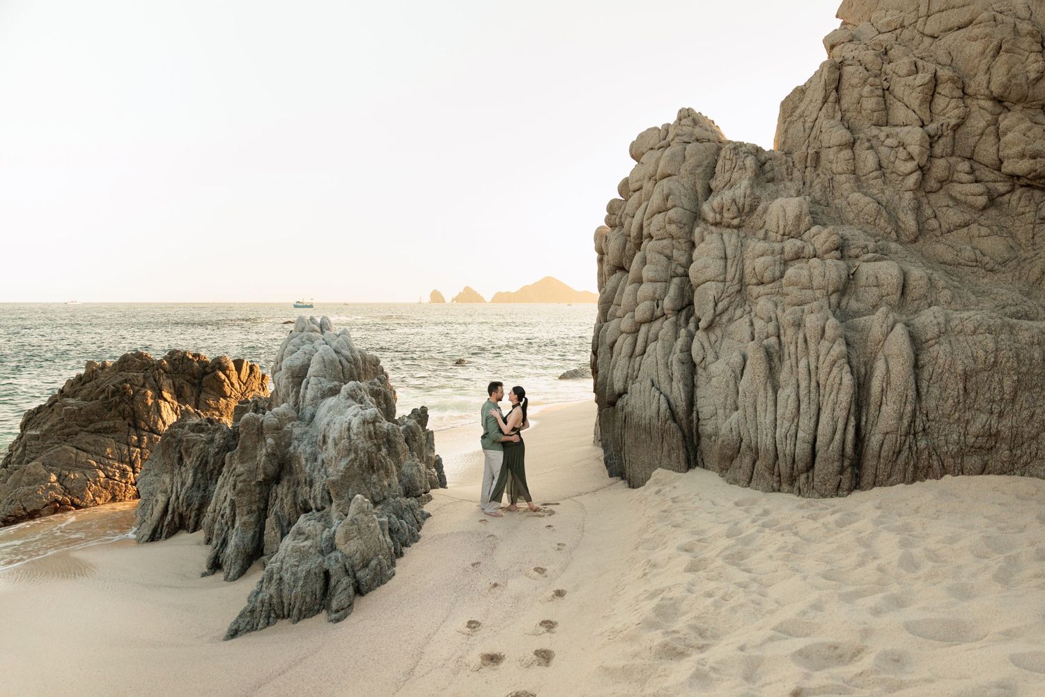 Couple embracing on sandy beach surrounded by dramatic rock formations at sunset in Cabo.
