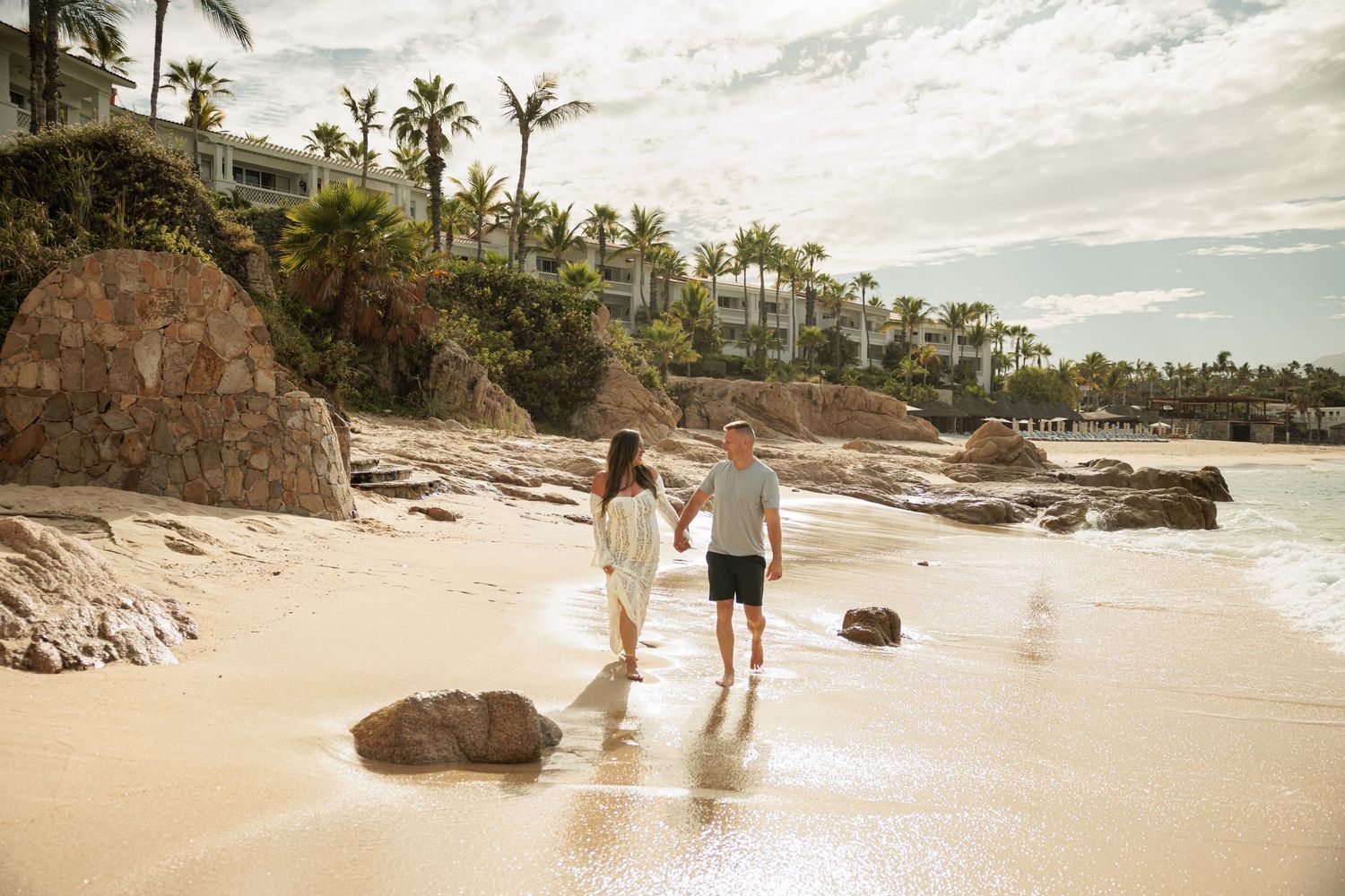 Couple holding hands walking on a sandy beach resort shoreline with palm trees and rocky cliffs.