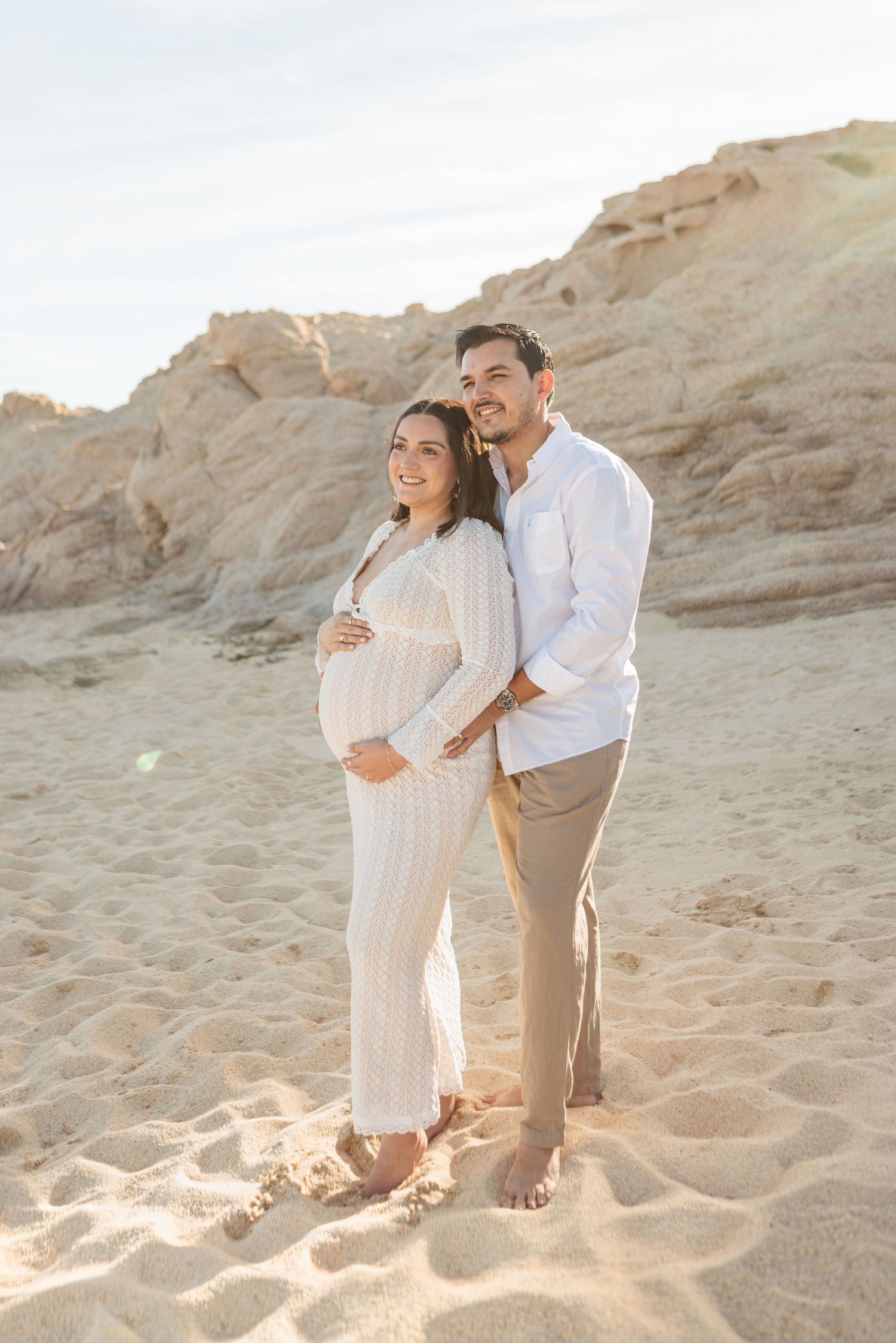 Pregnant couple posing on sandy beach with rocky cliffs, woman in white dress cradling baby bump, man in white shirt embracing her.