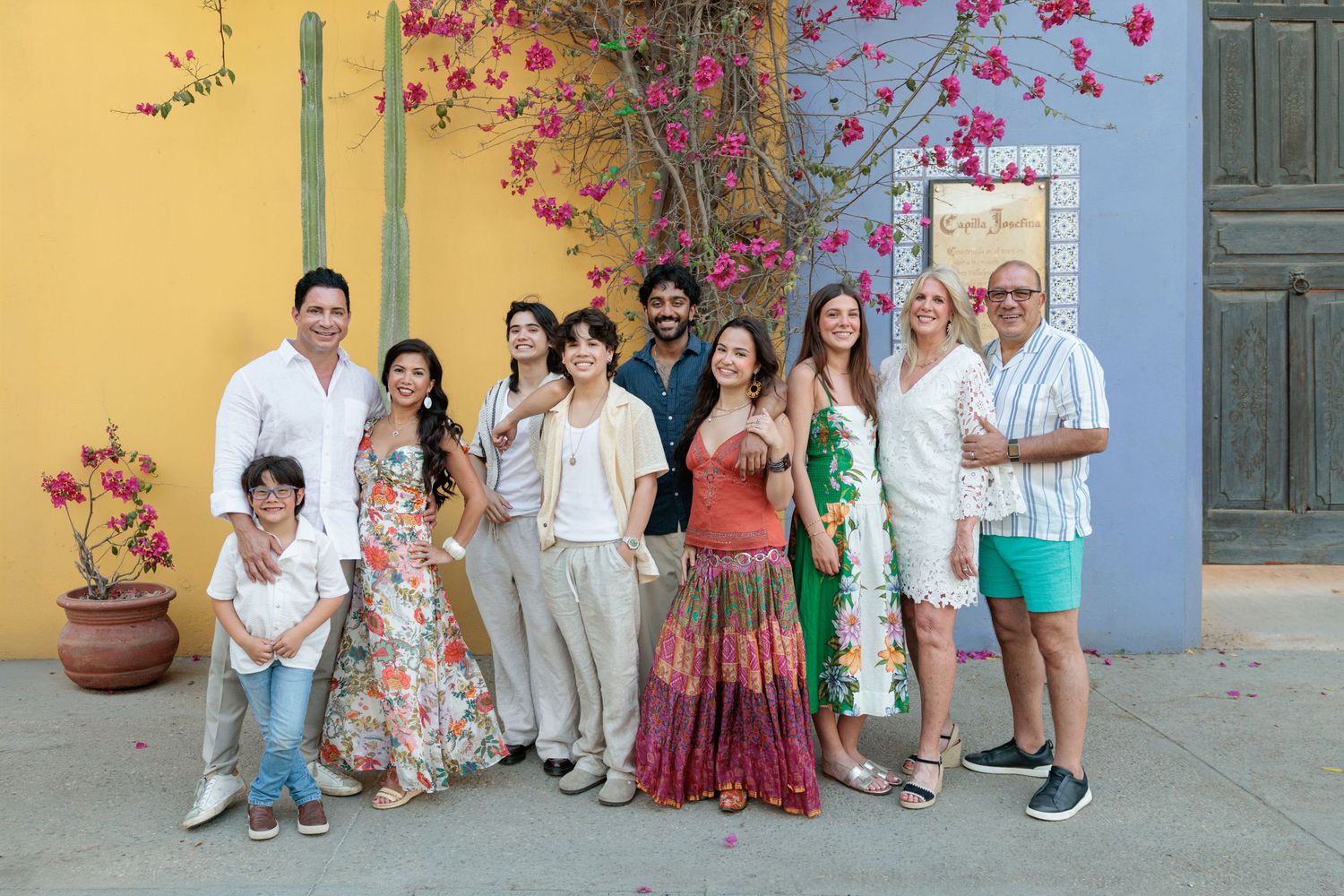 A diverse group of smiling people posing together in front of a colorful yellow and pink bougainvillea-adorned wall.