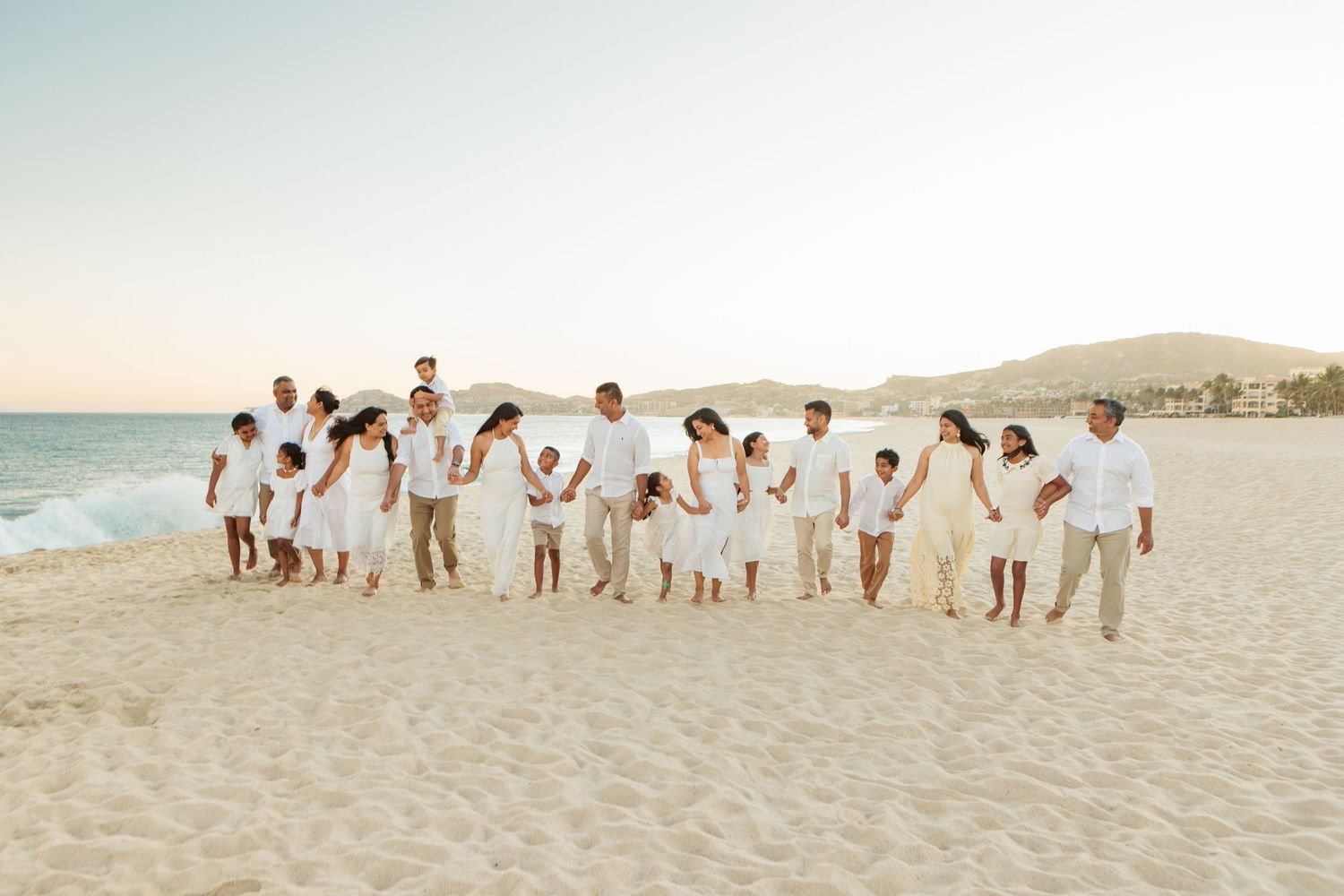 Large family in white outfits posing together on a sunny beach with ocean waves and mountains in the background.