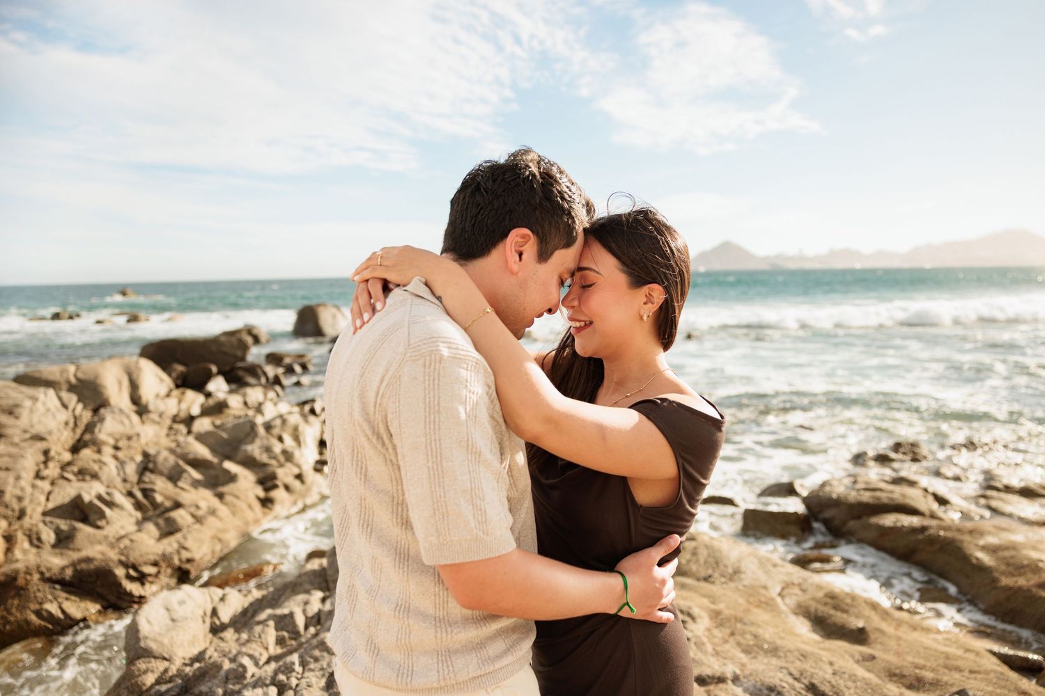 Romantic couple embracing forehead to forehead on a rocky oceanside beach under blue skies.