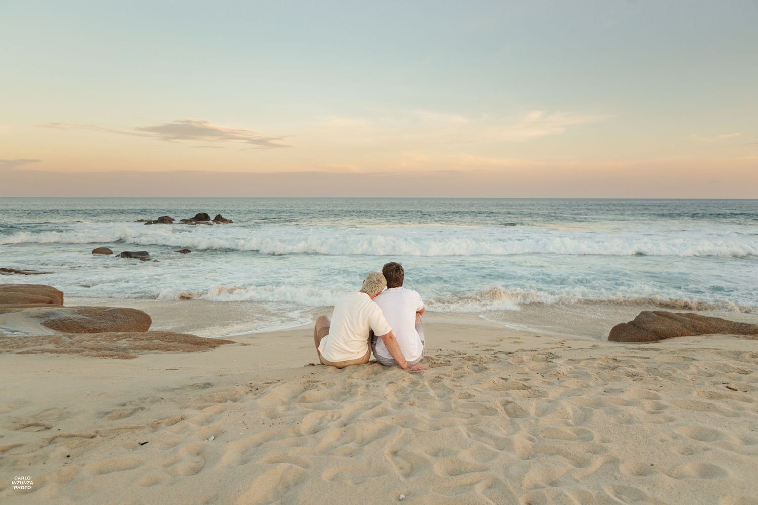 Couple sitting together on sandy beach facing ocean waves at golden sunset.