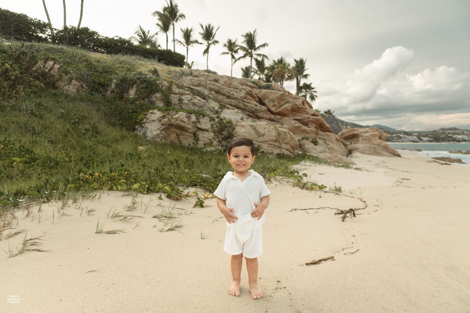 Adorable toddler in white outfit standing alone on sandy beach with rocky cliffs and palm trees.