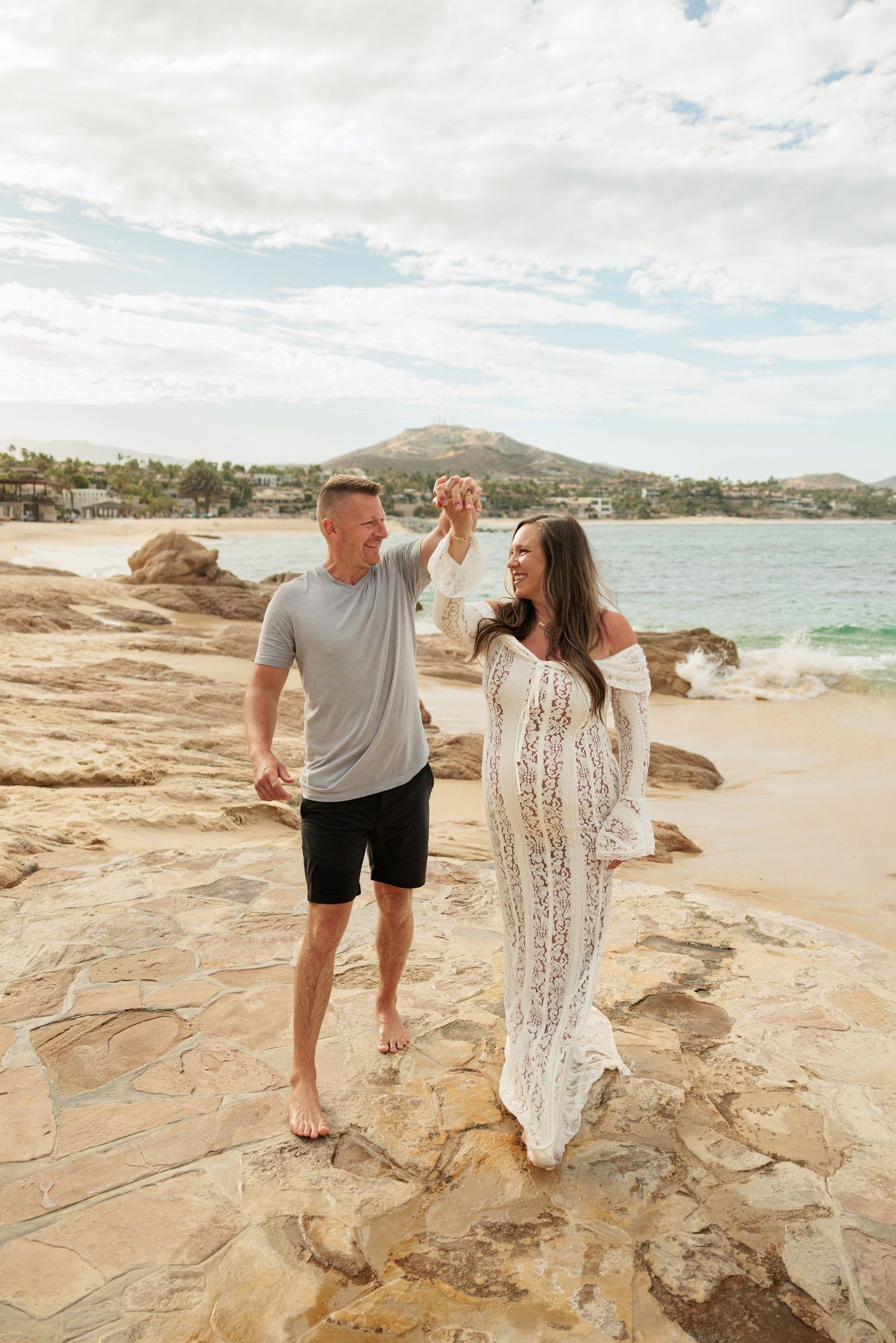 Couple dancing on rocky beach, woman in white lace dress, man in gray tee, ocean and mountain backdrop.