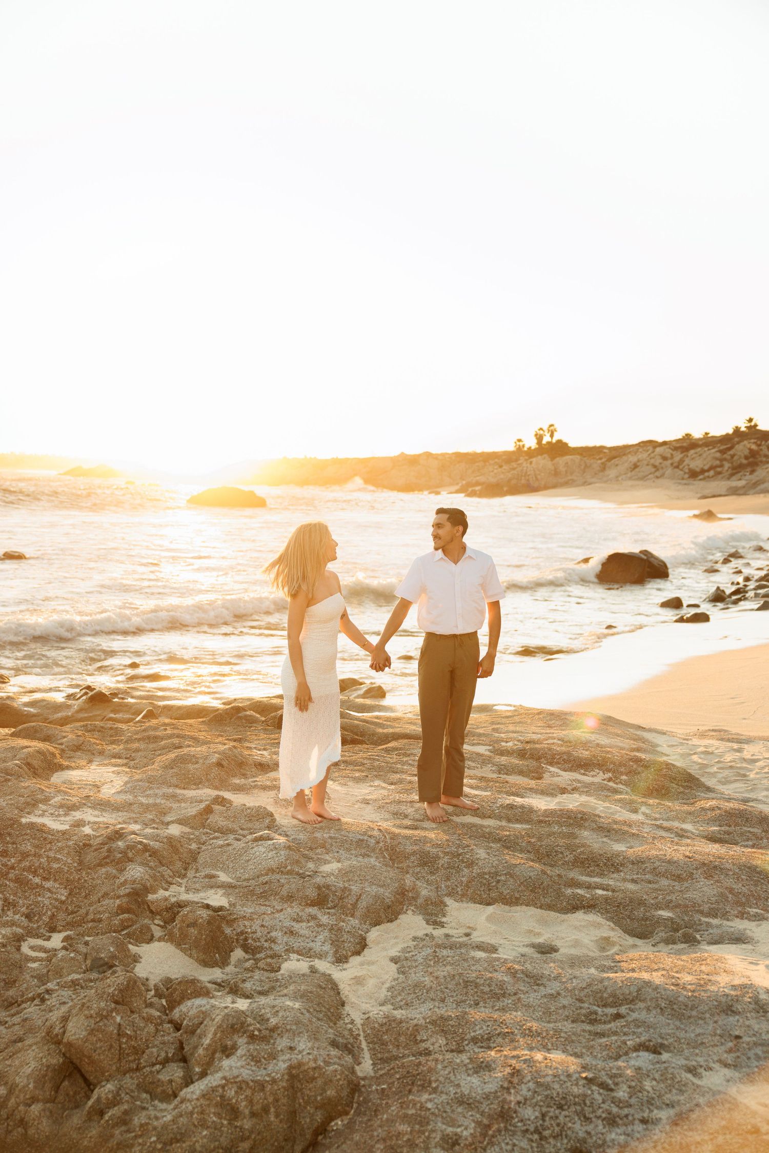 Couple holding hands on rocky beach at golden sunset, woman in white dress, man in white shirt and brown pants.