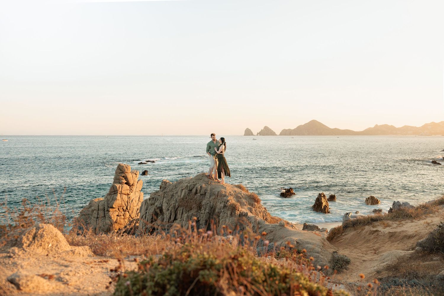 Couple embracing on rocky coastal cliffs at golden hour with ocean and mountains in background.