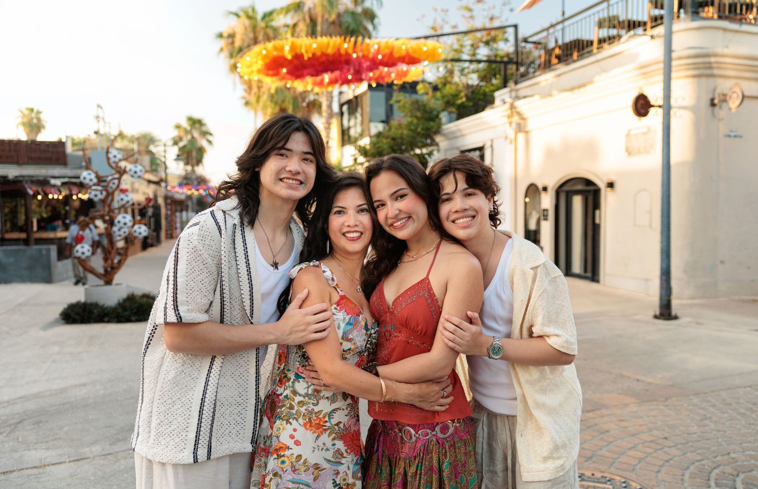 Four smiling friends pose together outdoors at a festive venue with colorful decorations on a sunny day.