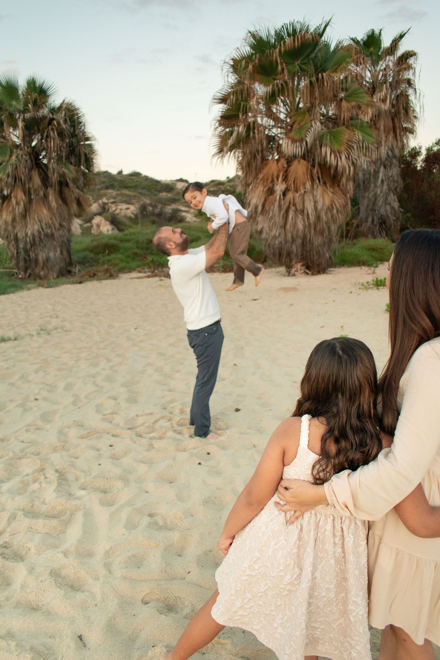 Father lifting young son in air on beach while mother and daughter watch nearby.