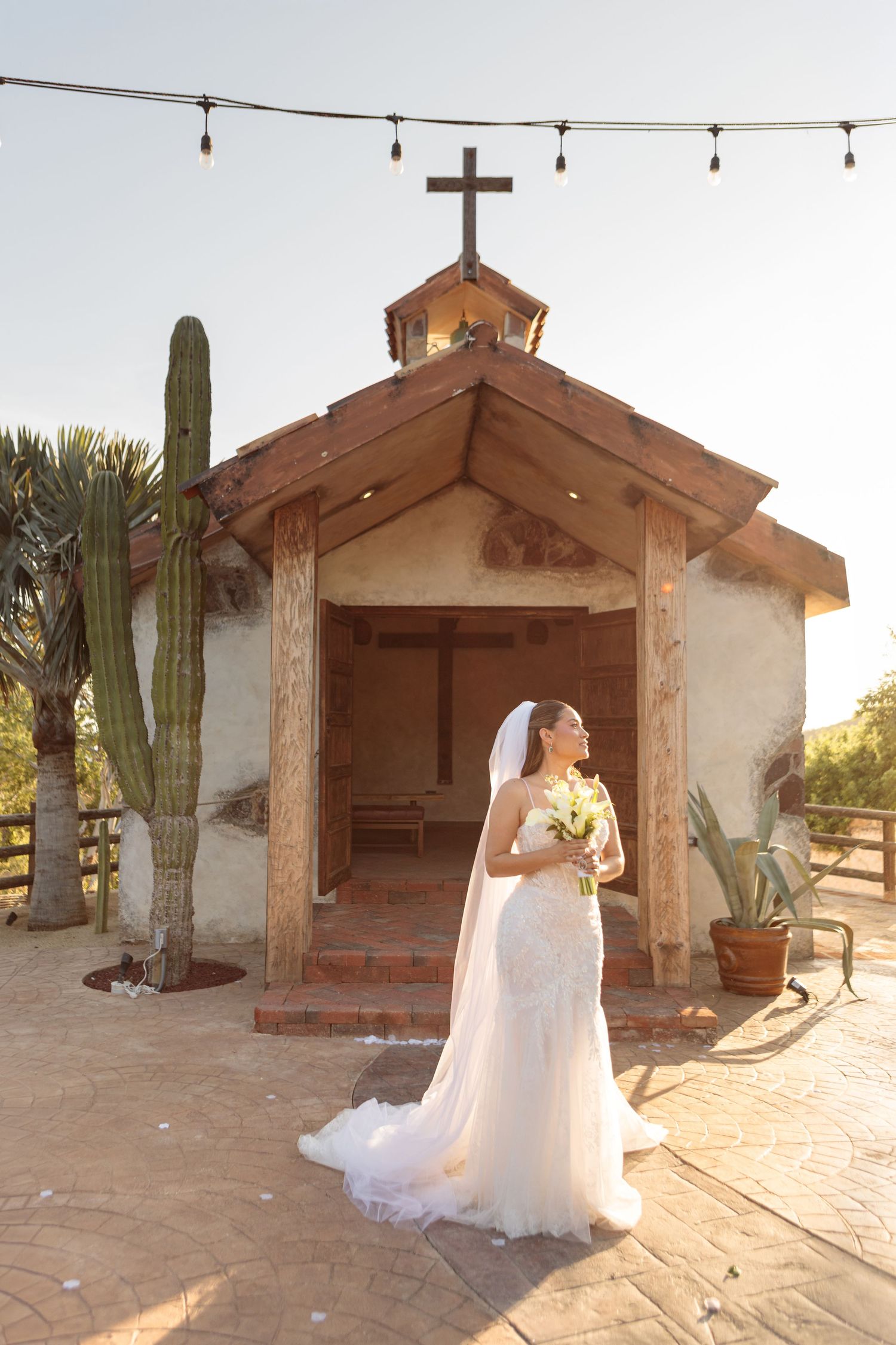 Elegant bride in lace mermaid gown holding bouquet outside rustic desert chapel at golden hour.