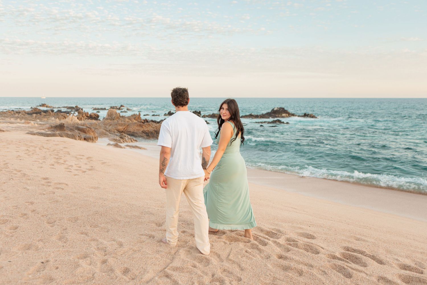 Couple holding hands walking toward ocean on a sandy beach at golden hour.