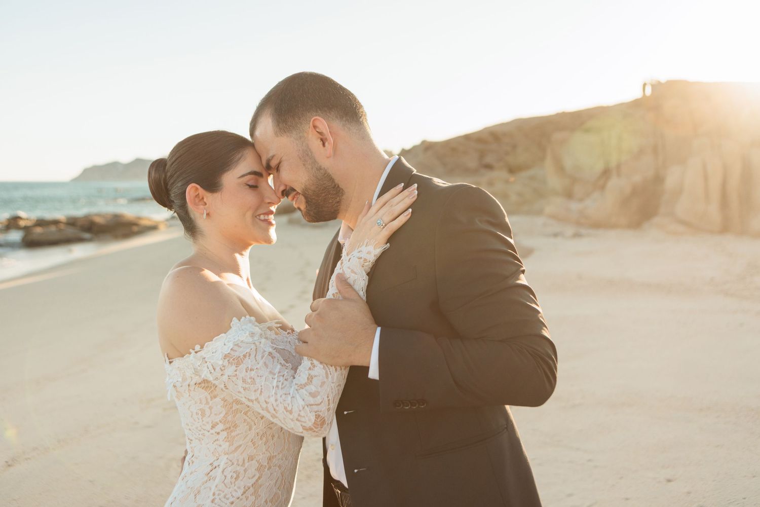 Bride and groom share a joyful embrace on sunlit beach, golden cliffs glowing behind them at sunset.