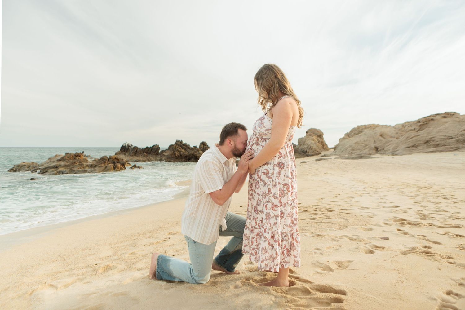 Man kneels on sandy beach kissing pregnant womans belly while she stands in floral dress by ocean.