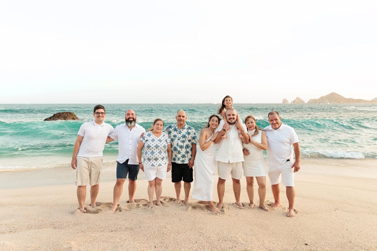 Group of nine people smiling on a sandy beach with turquoise ocean waves and rocky cliffs in the background.
