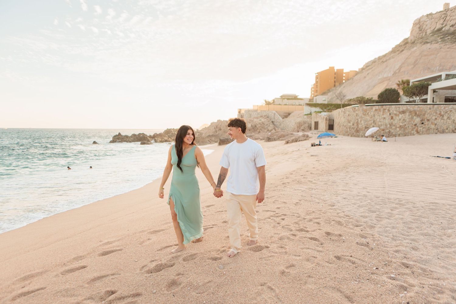 Couple holding hands walking along a sandy beach at golden hour with rocky cliffs in the background.