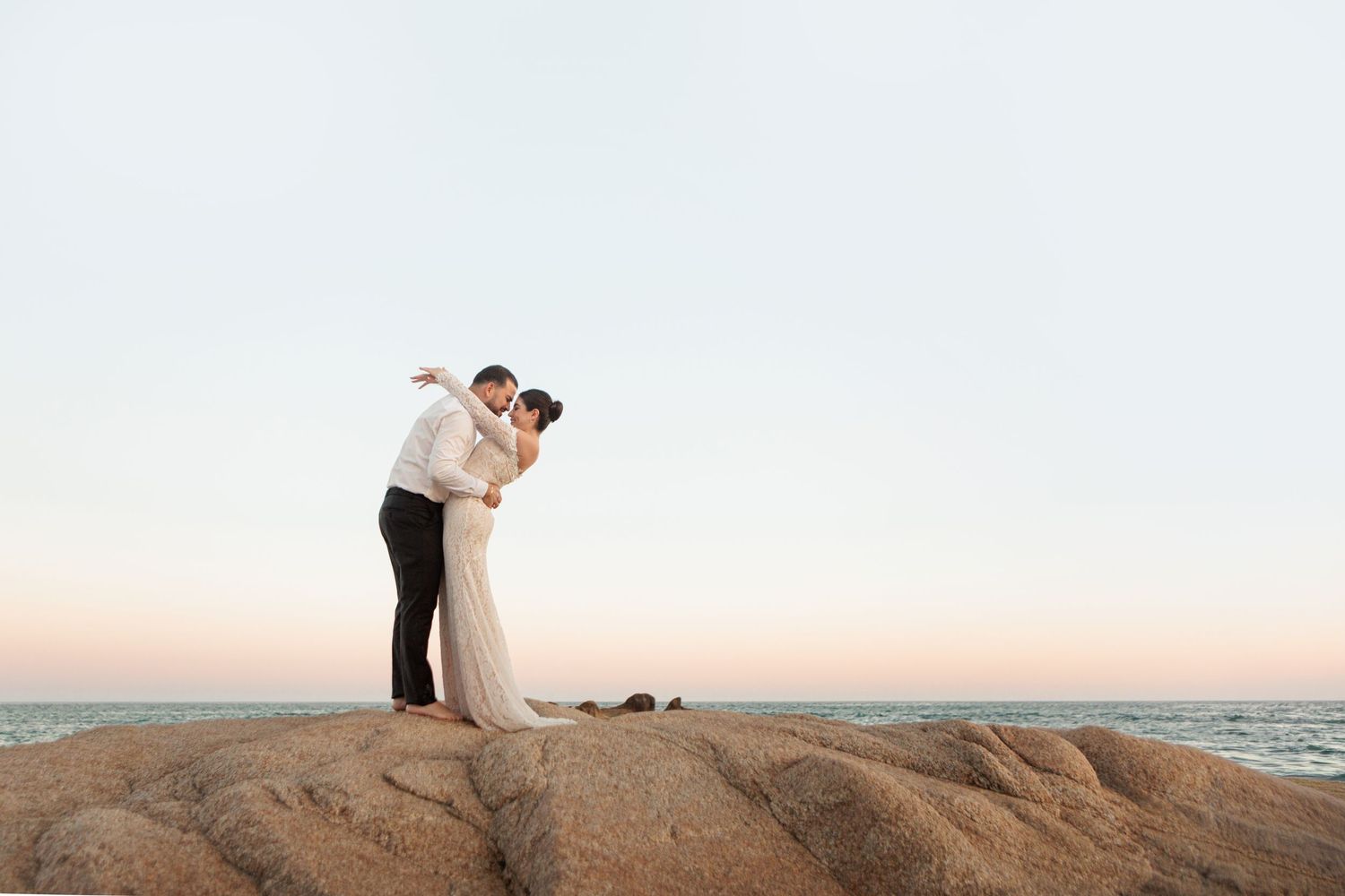 Bride and groom sharing a romantic kiss on rocky coastal cliffs at sunset during their beach wedding.