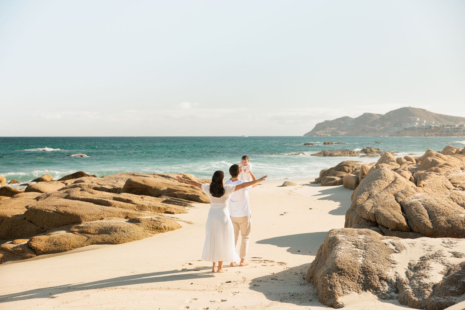 Couple in white outfits walking on a sandy beach with turquoise ocean waves and rocky shoreline.