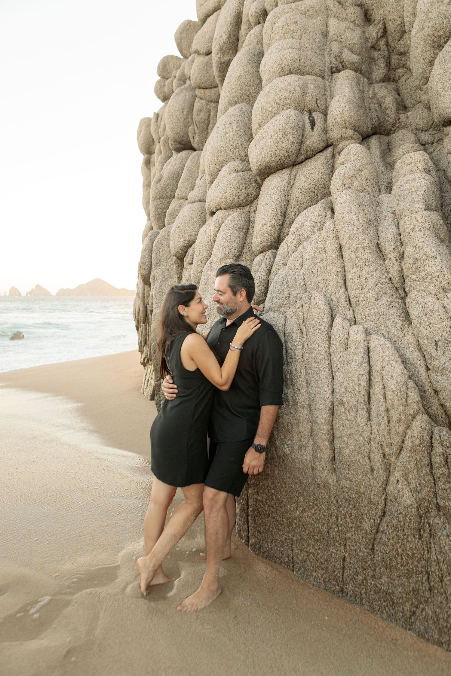 Elegant beach portrait of a couple embracing by the rock formations at the shore in Los Cabos.