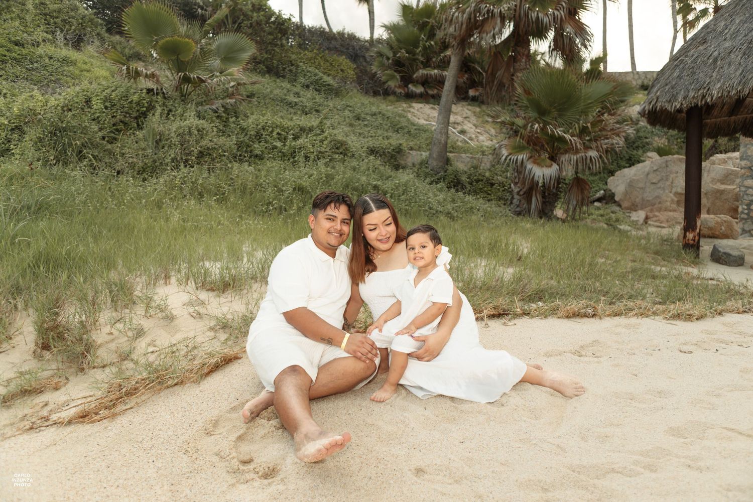 Family of three in white outfits sitting together on sand dunes surrounded by tropical greenery.