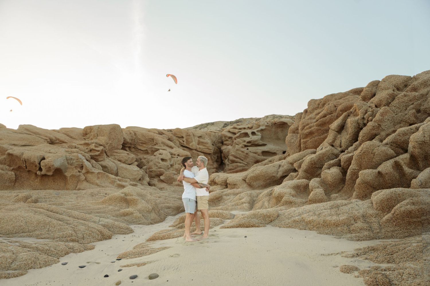 Couple embracing on sandy beach surrounded by eroded rock formations with paragliders overhead.