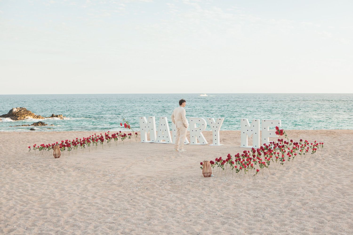 Romantic beach proposal setup with MARRY ME marquee letters and rows of red roses.